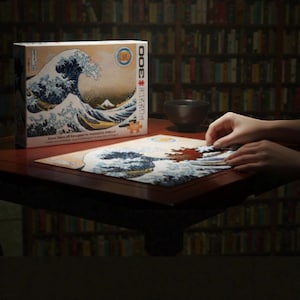 Peut inclure Un puzzle de 3000 pièces représentant une œuvre d'art japonaise célèbre, La Grande Vague de Kanagawa de Katsushika Hokusai. L'image représente une grande vague bleue qui se brise sur trois bateaux avec le mont Fuji en arrière-plan.