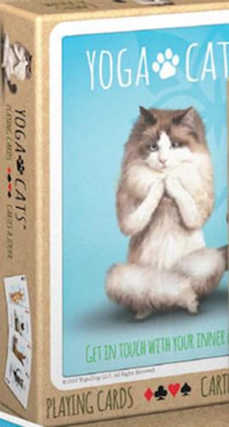 May include: A deck of playing cards featuring a white cat in a yoga pose. The box has a blue background with the text "Yoga Cats" and "Get in touch with your inner cat".