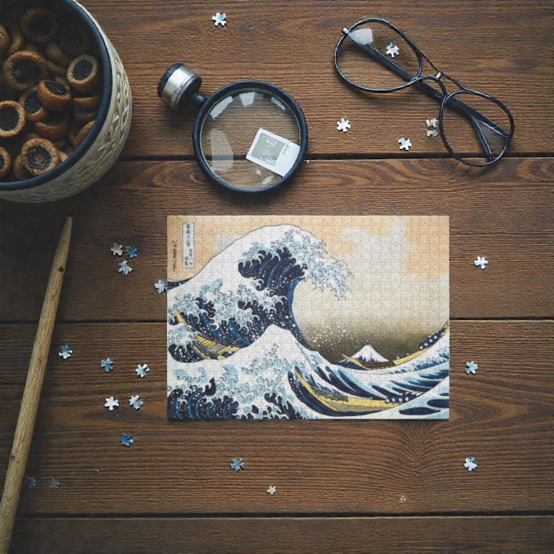 Peut inclure Un puzzle de 3000 pièces représentant une œuvre d'art japonaise célèbre, La Grande Vague de Kanagawa de Katsushika Hokusai. L'image représente une grande vague bleue qui se brise sur trois bateaux avec le mont Fuji en arrière-plan.