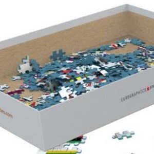 K&ouml;nnte beinhalten: Eine wei&szlig;e Box mit braunem Innenraum, gef&uuml;llt mit blauen, roten und gelben Puzzle-Teilen. Die Box tr&auml;gt den Text "EUROGRAPHICS PUZZLES" und eine Website-Adresse, die auf der Seite aufgedruckt ist.