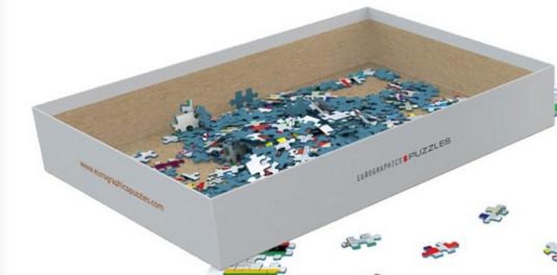 K&ouml;nnte beinhalten: Eine wei&szlig;e Box mit braunem Innenraum, gef&uuml;llt mit blauen, roten und gelben Puzzle-Teilen. Die Box tr&auml;gt den Text "EUROGRAPHICS PUZZLES" und eine Website-Adresse, die auf der Seite aufgedruckt ist.