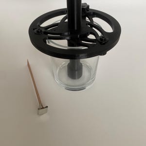 Puede incluir: Un portavelas negro impreso en 3D con un tarro de vidrio y un sumergidor de mecha de metal. El portavelas tiene una base circular con un diseño decorativo y un poste central que sostiene el tarro.