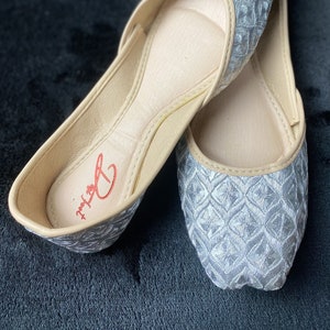 May include: A pair of silver embroidered flat shoes with a beige lining. The shoes have a geometric pattern and the brand name "Delux" is visible on the insole.