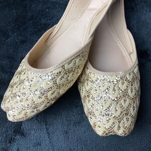 May include: A pair of gold embellished flat shoes with a pointed toe. The shoes have a delicate lace pattern and are made of a soft fabric. The brand name "The Pearl" is visible on the inside of one shoe.