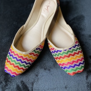 May include: A pair of colorful flat shoes with a zig-zag pattern in shades of red, orange, yellow, green, blue, and purple. The shoes have a gold thread woven into the fabric and a white leather insole.