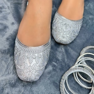 May include: A pair of silver embroidered flat shoes with a silver bangle bracelet set on a grey fuzzy surface.