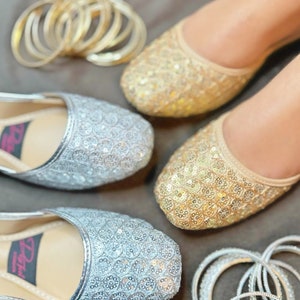 May include: Two pairs of flat shoes with gold and silver sequins. The shoes are decorated with small round sequins. The shoes are surrounded by gold and silver bangles.