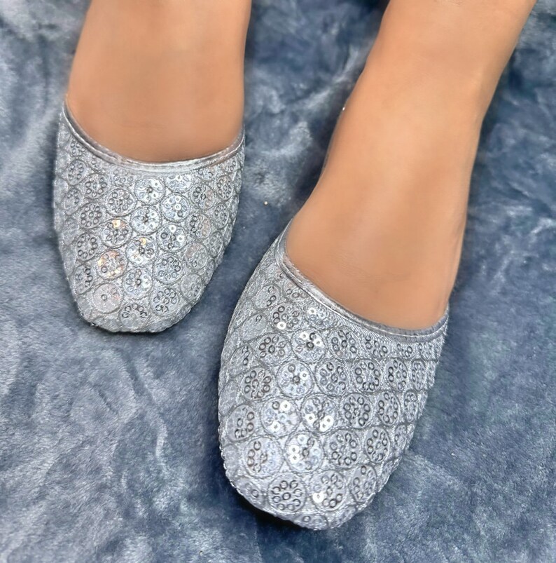 May include: A pair of silver sequined flat shoes. The shoes have a pointed toe and are decorated with small, round sequins.
