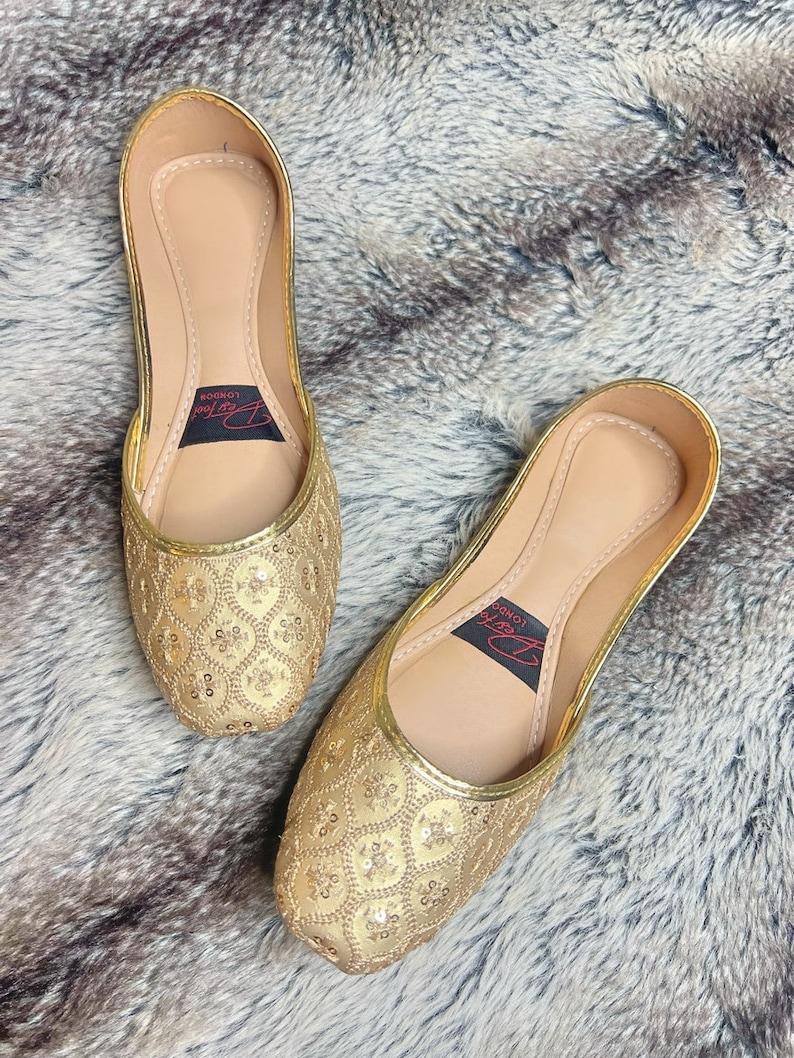K&ouml;nnte beinhalten: Ein Paar flache goldene Schuhe mit spitzen Zehen, verziert mit Pailletten. Die Schuhe haben ein Obermaterial aus goldenem Leder mit einem zarten Muster aus Pailletten. Der Markenname "London Look" ist auf der Innenseite der Schuhe aufgedruckt.