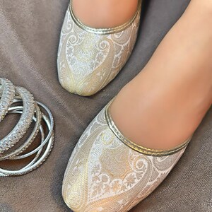 Op de afbeelding: Een paar platte, witte en gouden geborduurde schoenen met een spitse neus. De schoenen hebben een paisley-patroon en een gouden rand.