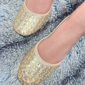 May include: A pair of gold sequined flat shoes with a pointed toe. The shoes are made of a soft, beige fabric and are decorated with small, round sequins.