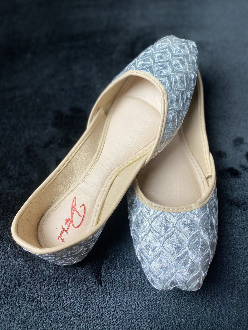 May include: A pair of silver embroidered flat shoes with a beige lining. The shoes have a geometric pattern and the brand name "Delux" is visible on the insole.