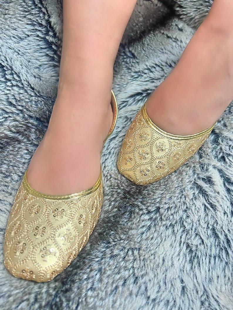 K&ouml;nnte beinhalten: Ein Paar flache goldene Schuhe mit Pailletten und spitzer Zehenpartie. Die Schuhe haben ein zartes Blumenmuster und sind aus einem weichen, bequemen Material gefertigt.