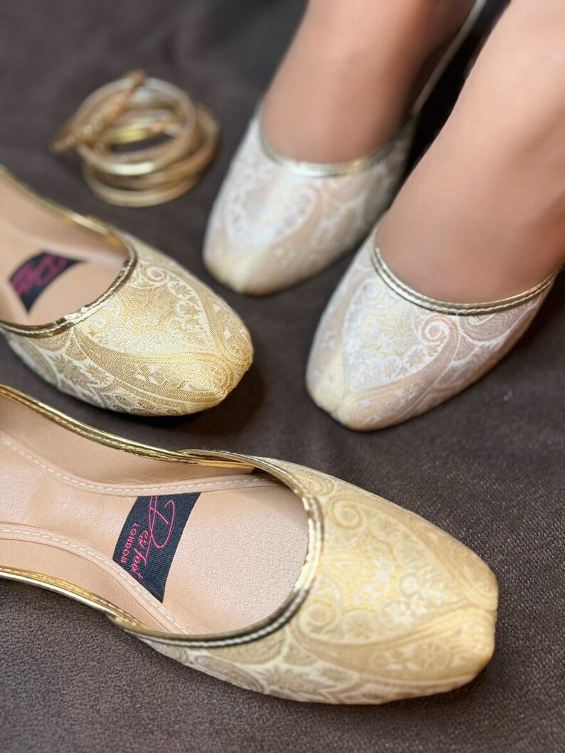 Op de afbeelding: Een paar platte, goud- en wit geborduurde schoenen met een spitse neus. De schoenen hebben een gouden rand en een zwart label met de tekst "Dil Tad London" op de binnenzool.