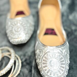 May include: A pair of silver embroidered flat shoes with a sparkly design. The shoes have a pointed toe and a low heel.