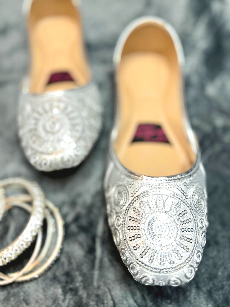 May include: A pair of silver embroidered flat shoes with a sparkly design. The shoes have a pointed toe and a low heel.