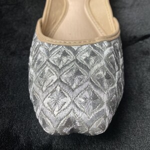 May include: Silver embroidered flat shoes with a geometric pattern. The shoes have a pointed toe and a low heel.