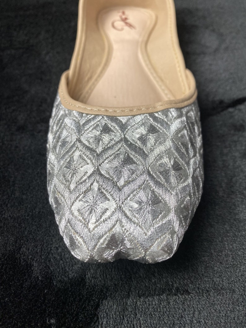 May include: Silver embroidered flat shoes with a geometric pattern. The shoes have a pointed toe and a low heel.
