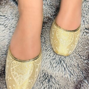 Op de afbeelding: Een paar platte gouden schoenen met een paisley-patroon. De schoenen zijn gemaakt van een zachte, comfortabele stof en hebben een spitse neus.