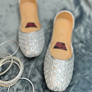 May include: A pair of silver sequined flat shoes with a pointed toe. The shoes have a decorative pattern of small circles and are made of a soft, comfortable material.