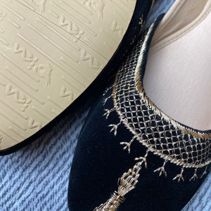 Op de afbeelding: Een paar zwarte su&egrave;de slippers met gouden borduurwerk. De slippers hebben een spitse neus en een platte zool. Het borduurwerk is ingewikkeld en heeft een geometrisch patroon.