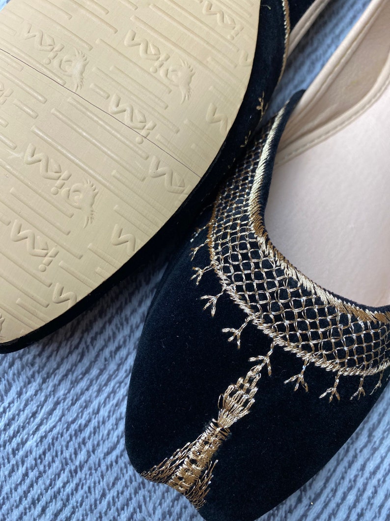 Op de afbeelding: Een paar zwarte su&egrave;de slippers met gouden borduurwerk. De slippers hebben een spitse neus en een platte zool. Het borduurwerk is ingewikkeld en heeft een geometrisch patroon.