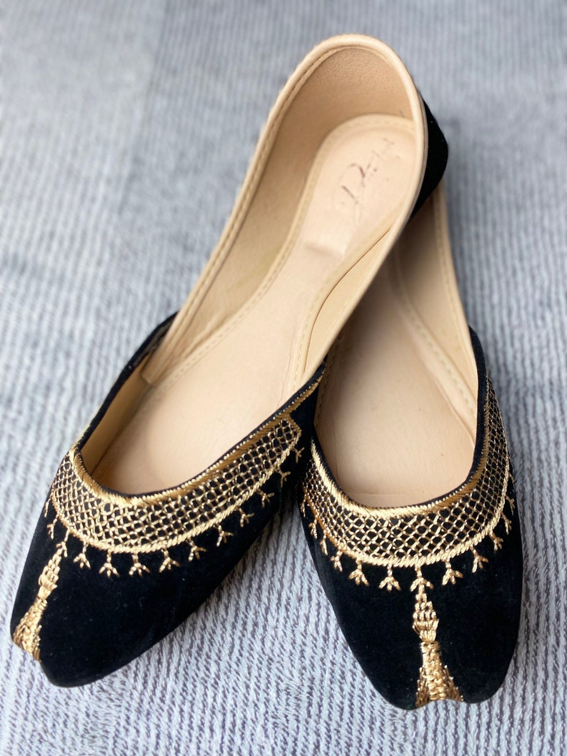 Op de afbeelding: Een paar zwarte fluwelen ballerina's met gouden borduurwerk en kwastjes. De schoenen hebben een spitse neus en een platte hak.
