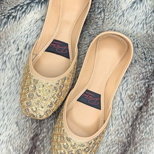 May include: A pair of gold embellished flat shoes with a beige leather upper. The shoes have a pointed toe and are decorated with gold sequins. The brand name "Art Foot London" is printed on the inside of the shoes.