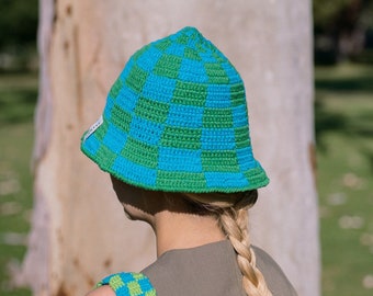 May include: A green and blue checkered bucket hat with a crocheted design. The hat is worn by a person with blonde hair.