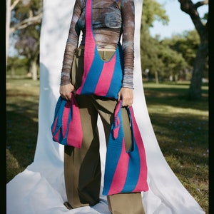 May include: Two pink and blue striped crocheted tote bags. The larger bag is worn over the shoulder and the smaller bag is held in the hand.