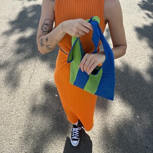 May include: A person wearing an orange ribbed sleeveless dress and black sneakers is holding a green and blue striped crocheted tote bag.