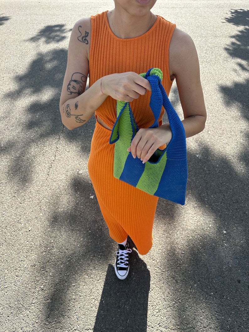 May include: A person wearing an orange ribbed sleeveless dress and black sneakers is holding a green and blue striped crocheted tote bag.