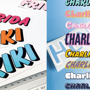 May include: A collection of colourful iron-on name labels with different fonts and styles. The names include "Frida", "Kiki", and "Charlie".