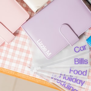 May include: Purple leather binder with the name "Lizzie M" printed on the front. The binder has clear plastic pockets labelled "Car", "Bills", "Food", "Holiday", and "Wedding".