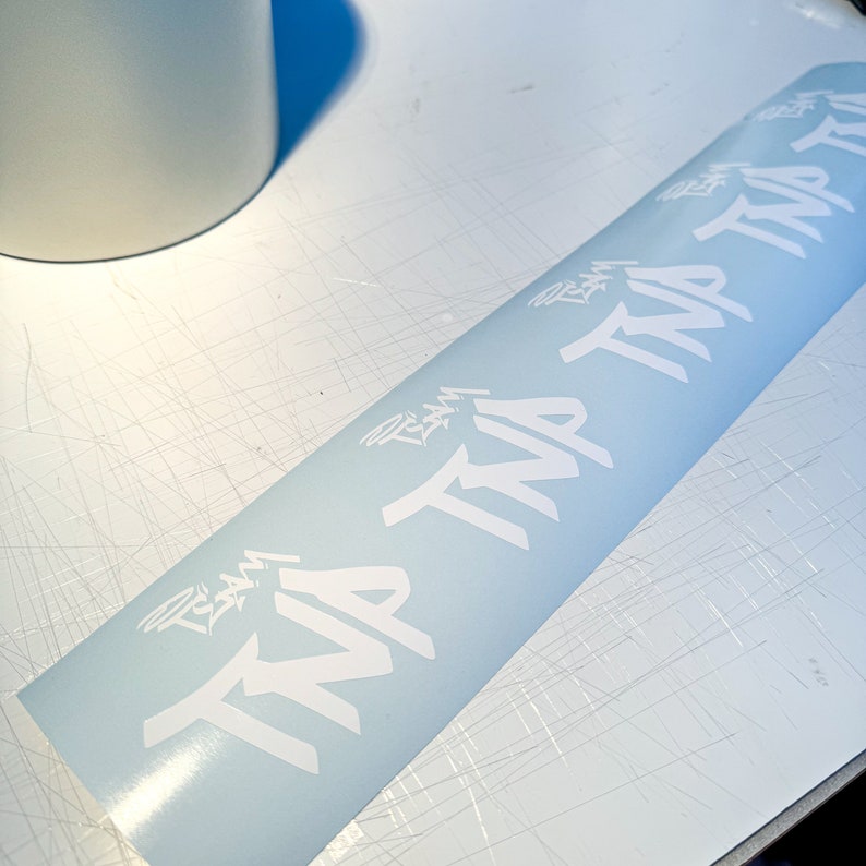 May include: A white vinyl sticker with the words "TRIA" repeated multiple times in a stylized font. The sticker is on a light blue background.