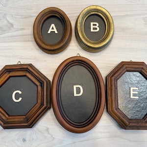 May include: Five vintage wooden picture frames in various shapes. Two oval frames, one with a gold finish, and three octagonal frames. Each frame has a dark interior and a letter in the center: A, B, C, D, and E.