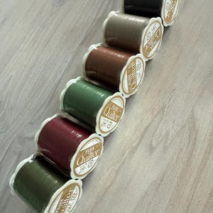 May include: An assortment of seven spools of quilting thread in various colors, including black, tan, brown, green, burgundy, and olive green. Each spool is labeled with the brand name "Fujix Quilter" in a white and gold label.