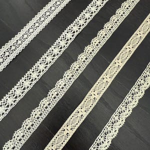 May include: Several strips of off-white lace with various openwork patterns are arranged diagonally against a dark background. The lace designs include floral and geometric motifs, showcasing the delicate textures and details.