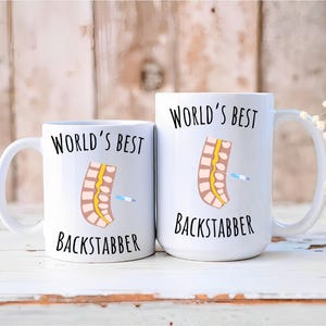 May include: Two white ceramic mugs with black text and a cartoon image of a spine. The text on the mugs reads "World's Best Backstabber".