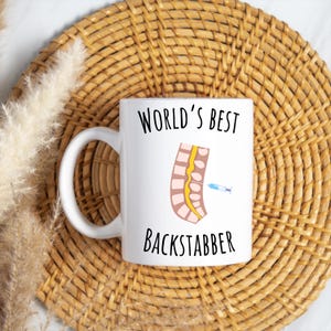 11 oz white glossy mug, Worlds Best Backstabber in black font, picture of blue and gray needle going into the spine, spine is tan color, spinal cord is yellow, depicting the spinal anesthesia process