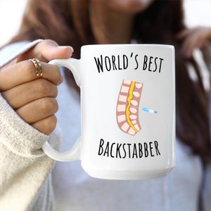 15 oz white glossy mug, Worlds Best Backstabber in black font, picture of blue and gray needle going into the spine, spine is tan color, spinal cord is yellow, depicting the spinal anesthesia process