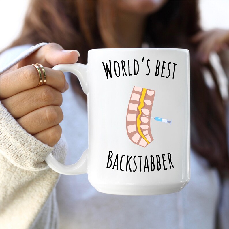 15 oz white glossy mug, Worlds Best Backstabber in black font, picture of blue and gray needle going into the spine, spine is tan color, spinal cord is yellow, depicting the spinal anesthesia process