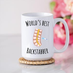 15 oz white glossy mug, Worlds Best Backstabber in black font, picture of blue and gray needle going into the spine, spine is tan color, spinal cord is yellow, depicting the spinal anesthesia process