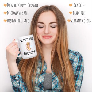 15 oz white glossy mug, Worlds Best Backstabber in black font, picture of blue and gray needle going into the spine, spine is tan color, spinal cord is yellow, depicting the spinal anesthesia process