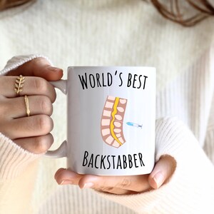 11 oz white glossy mug, Worlds Best Backstabber in black font, picture of blue and gray needle going into the spine, spine is tan color, spinal cord is yellow, depicting the spinal anesthesia process