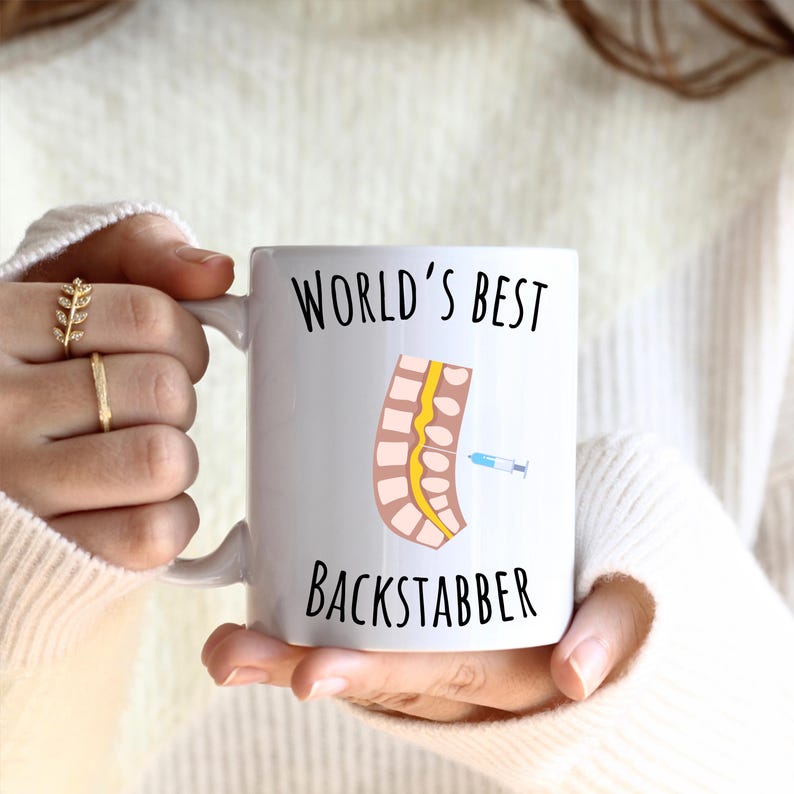 11 oz white glossy mug, Worlds Best Backstabber in black font, picture of blue and gray needle going into the spine, spine is tan color, spinal cord is yellow, depicting the spinal anesthesia process