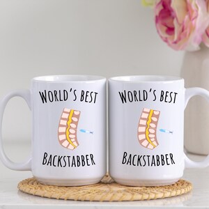 15 oz white glossy mugs, Worlds Best Backstabber in black font, picture of blue and gray needle going into the spine, spine is tan color, spinal cord is yellow, depicting the spinal anesthesia process