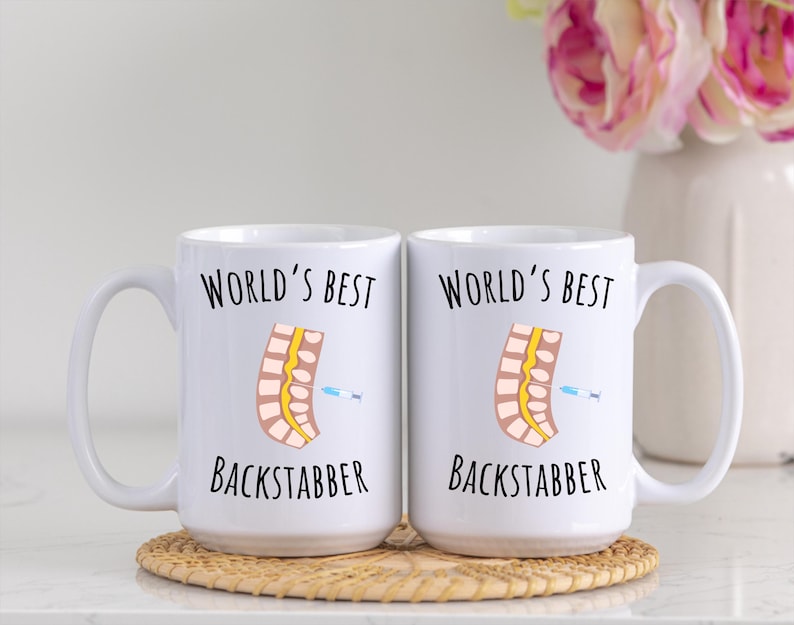 15 oz white glossy mugs, Worlds Best Backstabber in black font, picture of blue and gray needle going into the spine, spine is tan color, spinal cord is yellow, depicting the spinal anesthesia process