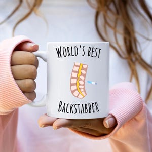 11 oz white glossy mug, Worlds Best Backstabber in black font, picture of blue and gray needle going into the spine, spine is tan color, spinal cord is yellow, depicting the spinal anesthesia process