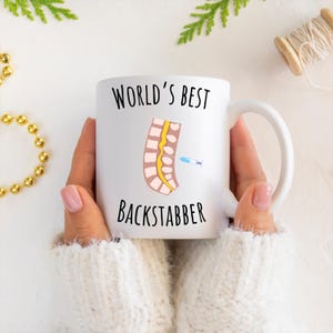 11 oz white glossy mug, Worlds Best Backstabber in black font, picture of blue and gray needle going into the spine, spine is tan color, spinal cord is yellow, depicting the spinal anesthesia process